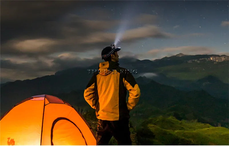 Camper wearing headlamp standing by orange tent in mountains at night