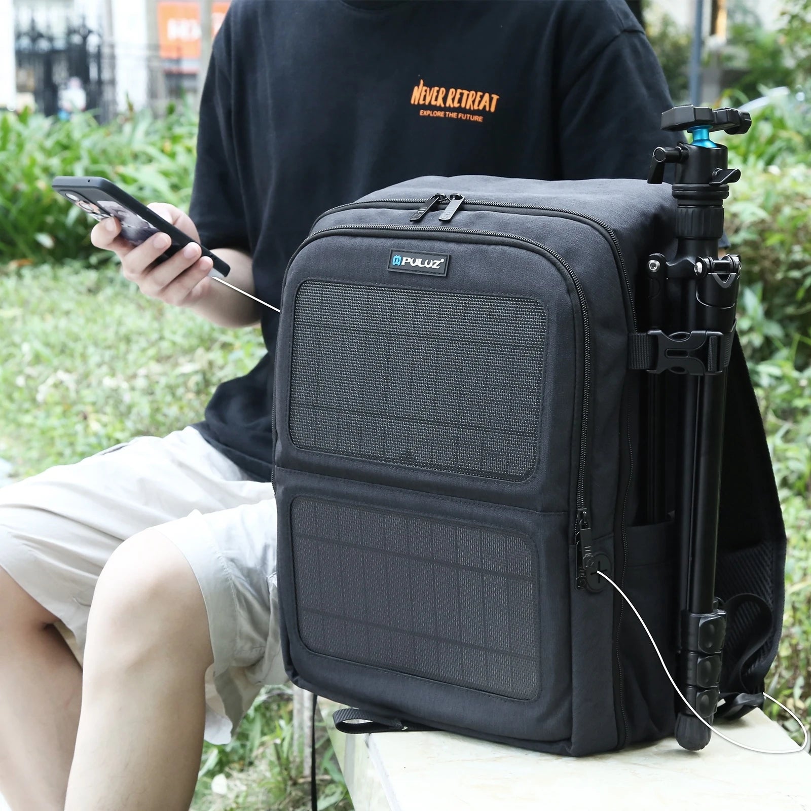 Outdoor backpack with solar panel charging a phone, tripod attached, person seated nearby.